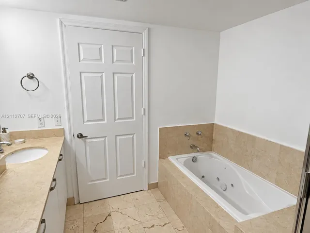 Jetted soaking tub with tile surround, stone-finish countertop, undermount sink, and neutral tile flooring