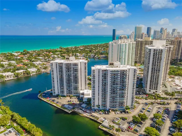 Aerial perspective of high-rise structures situated on a waterfront peninsula with direct coastal access and proximity to urban skyline, featuring on-site surface parking lots and established tropical landscaping