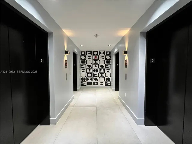 Hallway featuring large-format light-toned floor tiling, matte black door finishes, geometric decorative wall paneling, and wall-mounted cylindrical sconces