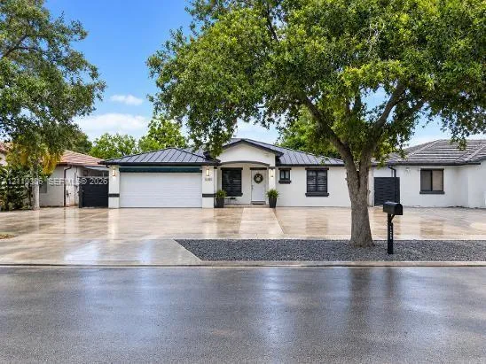 Single-story exterior featuring a standing-seam metal roof, stucco finish, attached two-car garage, concrete driveway, and mature landscaping