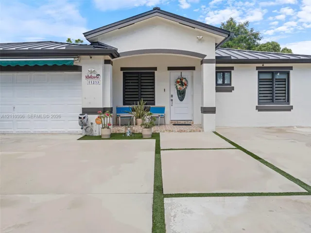 Exterior featuring a white stucco facade, metal roof, concrete driveway with synthetic grass inlay, covered entryway, and dark-framed windows