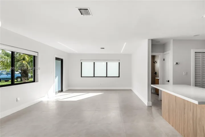 Open interior featuring large-format tile flooring, recessed linear lighting, expansive windows with black frames, and a kitchen island with a light-toned wood-finish base and quartz countertop