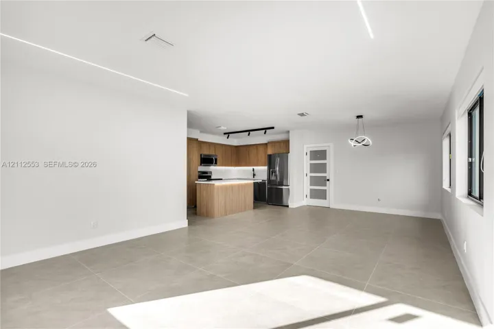 Open-concept interior featuring large-format tile flooring, recessed linear lighting, kitchen with wood-finish cabinetry, center island, and a multi-pane frosted glass door