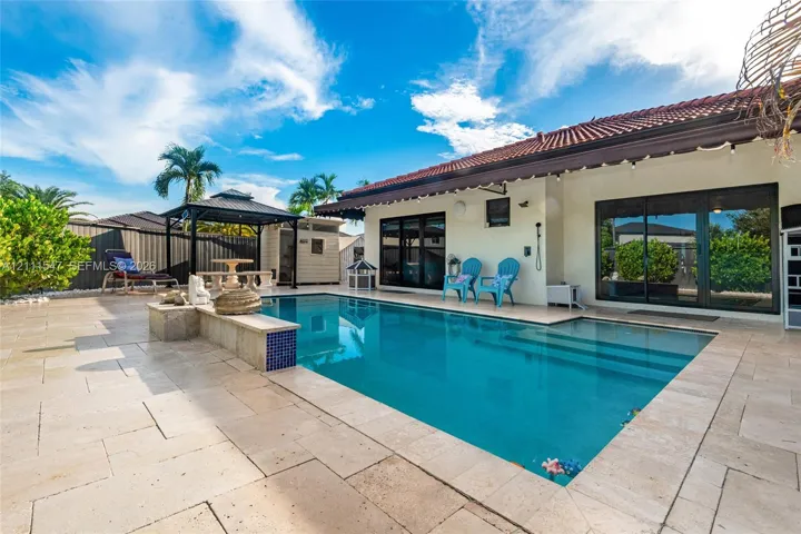 In-ground swimming pool featuring a tiled accent wall, stone-style deck surround, covered patio area, and detached metal-frame gazebo