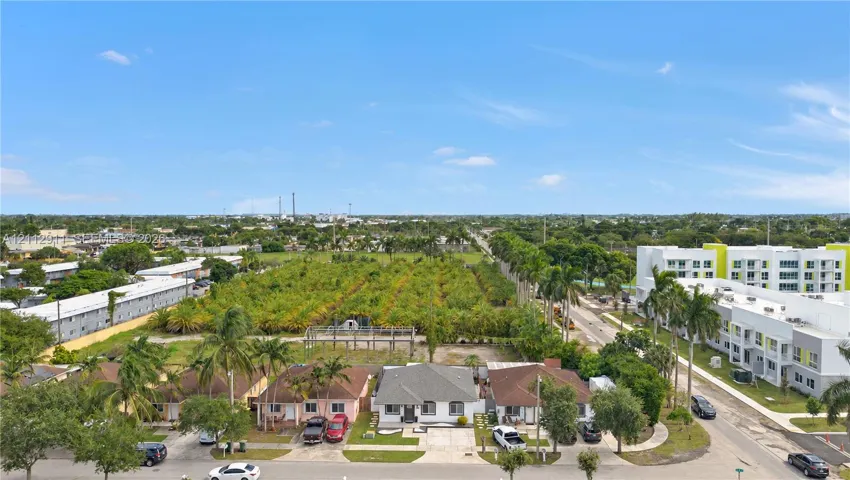 Aerial perspective featuring a single-story residence with a pitched roof, paved driveway, expansive level lot, and mature palm landscaping
