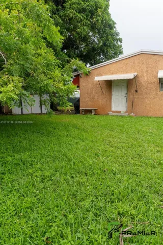Rear yard featuring a grass lawn, mature trees, textured stucco exterior, and a metal door awning