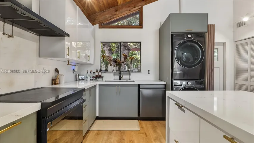 Modern kitchen featuring high-gloss white upper cabinetry, flat-panel base cabinets, quartz countertops, a vaulted wood-paneled ceiling, and a built-in laundry area