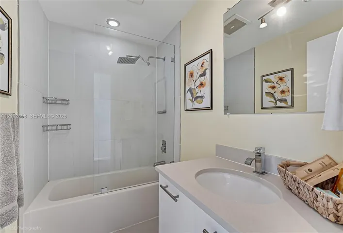 Bathroom featuring a vanity with a solid-surface countertop, rectangular undermount sink, and chrome faucet, paired with a glass-enclosed shower and tub combination featuring white wall tiling and a rainfall showerhead
