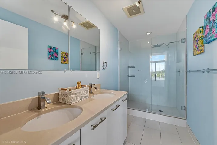 Dual-sink vanity with a solid-surface countertop, full-width mirror, glass-enclosed shower with a rainfall showerhead, and tile flooring