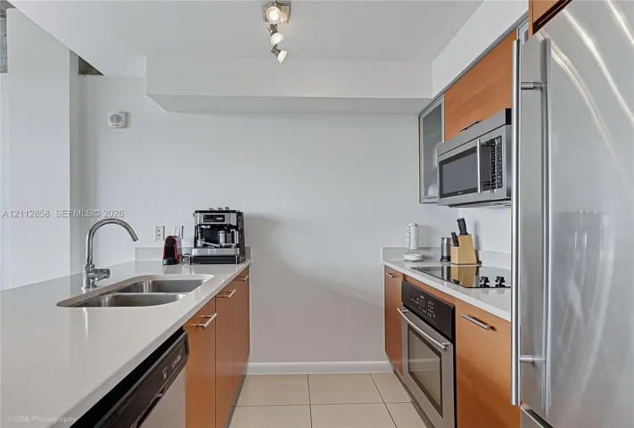Galley kitchen featuring flat-panel wood cabinetry, white stone-finish countertops, stainless steel appliances, dual-basin undermount sink, and tile flooring