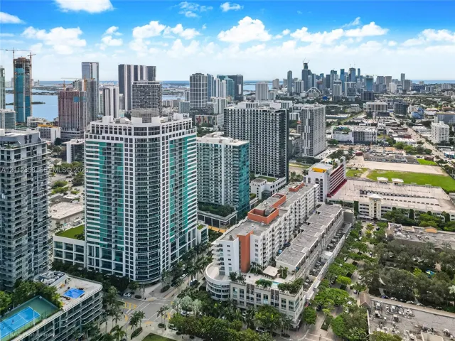 Aerial perspective of a high-rise urban environment featuring extensive glass-facade skyscrapers, multi-level parking structures, and established palm tree landscaping