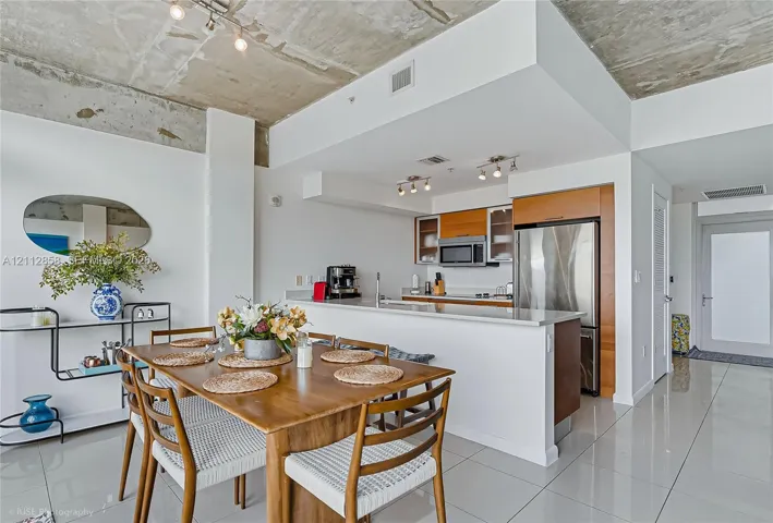 Open-concept kitchen featuring stainless steel appliances, wood-toned cabinetry, and a stone-surface breakfast bar under an exposed concrete ceiling with polished tile flooring