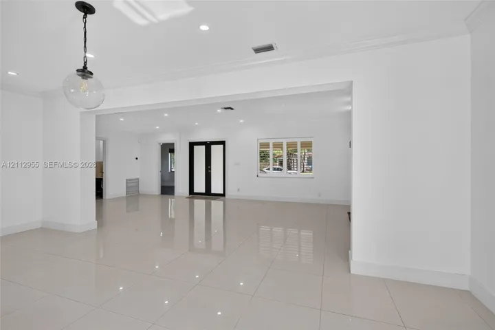 Open-concept interior featuring large-format porcelain tile flooring, crown molding, recessed lighting, double entry doors, and a decorative glass pendant light fixture