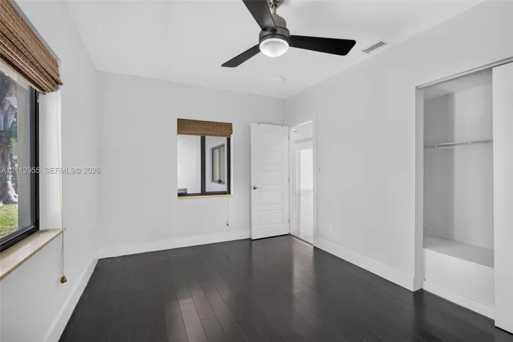 Room featuring dark wood-finish flooring, a contemporary matte black ceiling fan with integrated lighting, two windows with woven shades, and a built-in closet with sliding doors