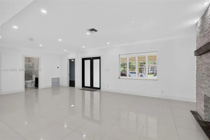 Large-format tile flooring, recessed ceiling lighting, stone-clad fireplace with a timber mantel, double-entry front door with glass insets, and windows featuring plantation shutters