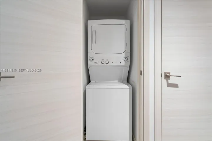 Laundry closet featuring a white stacked washer and dryer unit, light wood-finish doors, and brushed metallic hardware