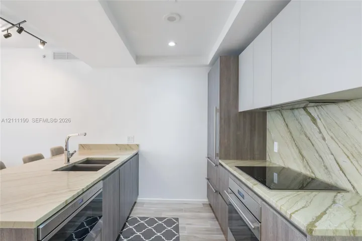Kitchen featuring stone countertops, stone backsplash, flat-panel wood-finish cabinetry, stainless steel sink with pull-down faucet, and built-in induction​​‌​​​​‌​​‌‌​‌‌​​​‌‌​‌​‌​‌​​​‌​​ cooktop