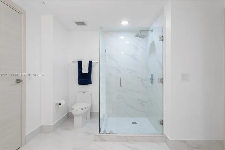 Walk-in glass-enclosed shower featuring stone-finish wall tile, dual-flush elongated toilet, and light-tone wood-finish door