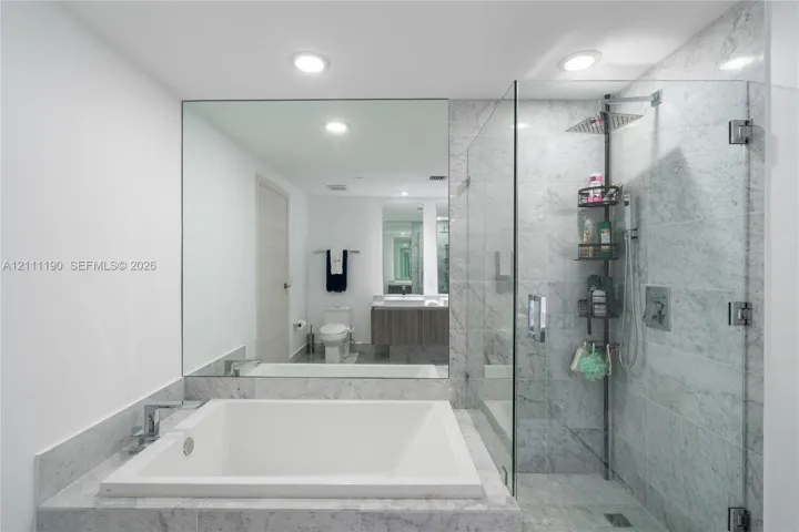Deep soaking tub with stone tile surround, frameless glass-enclosed shower, expansive wall-mounted mirror, recessed lighting, and stone flooring