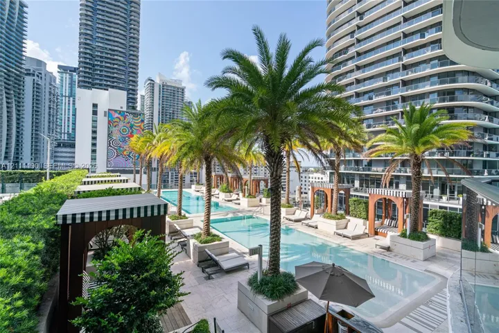 Elevated outdoor terrace featuring a multi-section swimming pool, stone tile decking, built-in wooden cabanas, and mature palm trees