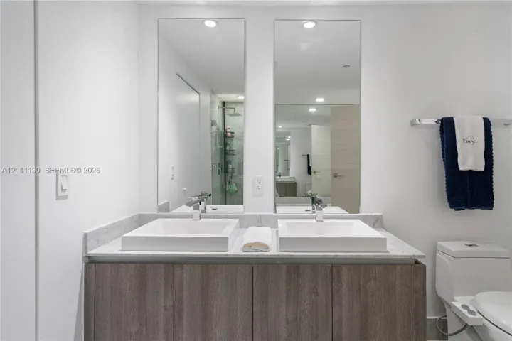 Dual-sink vanity featuring textured wood-finish cabinetry, a stone countertop, two rectangular vessel sinks, and wall-mounted vertical mirrors