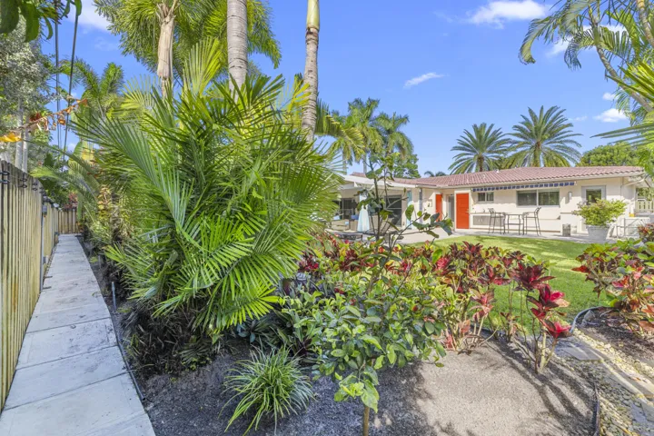 Rear exterior featuring a concrete walkway, mature palm trees, diverse tropical landscaping, and a fenced perimeter
