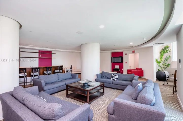 Open-concept interior featuring curved structural columns, recessed ceiling lighting, a built-in wet bar with wood-finish counter, and patterned carpet tiles