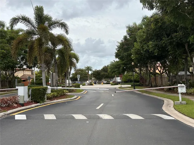 Gated community entrance featuring motorized security arms, asphalt roadway, concrete speed bumps, and mature palm and hardwood landscaping