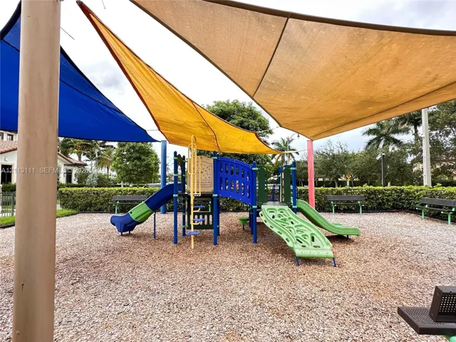 Outdoor recreational area featuring multiple shade sails, a multi-component play structure with dual slides, mulch ground cover, and metal bench seating