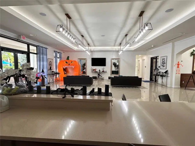 Open-concept room featuring light-colored floor tiling, recessed tray ceilings, four modern rectangular pendant light fixtures, and a large stone-surface countertop