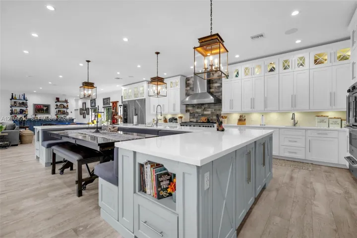 Kitchen featuring shaker-style cabinetry with glass-panel upper doors, a large central island with integrated storage, white quartz countertops, wood-finish flooring, and a stone-tile backsplash behind a stainless steel range hood