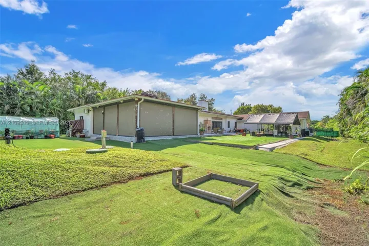 Expansive backyard featuring artificial turf, a permanent greenhouse structure, a covered patio with integrated privacy screens, and a timber-framed garden bed