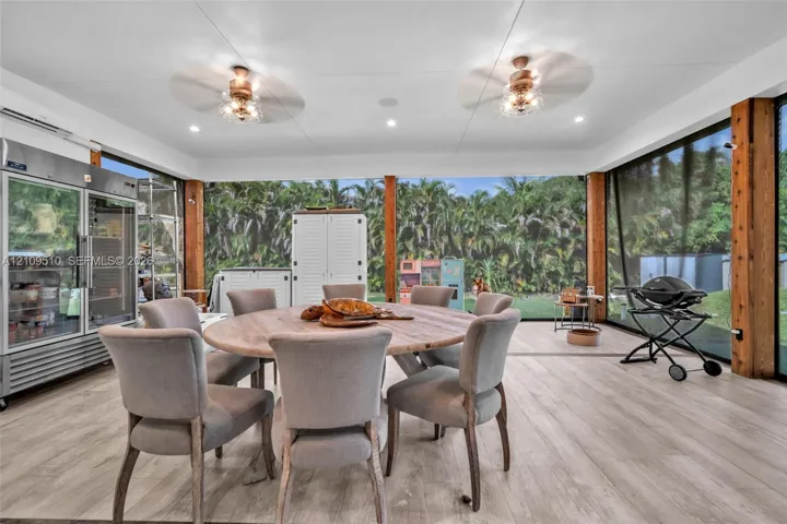 Enclosed outdoor living space featuring wood-finish flooring, exposed wood support pillars, dual ceiling fans, and an integrated stainless steel glass-door refrigerator