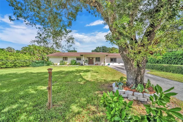 Single-story exterior featuring a light-toned stucco façade, a multi-paneled front entry door, expansive lawn, mature tree, and a concrete paver retaining wall