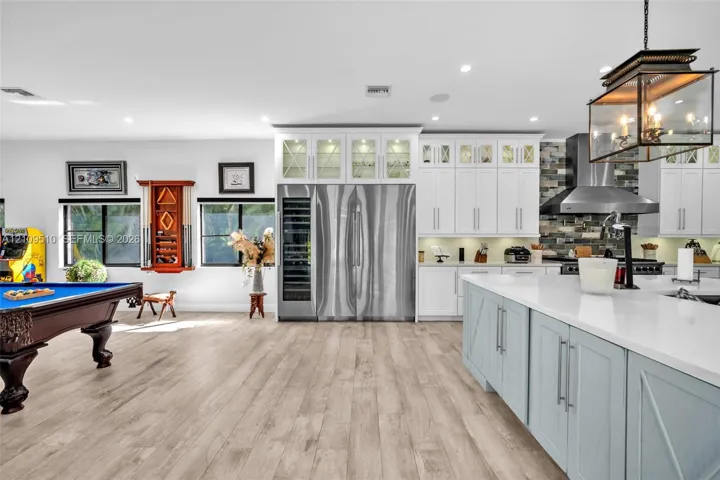 Kitchen featuring custom white cabinetry with glass-front uppers, a stainless steel side-by-side refrigerator, a built-in wine cooler, a stone tile backsplash, and a central island with a quartz countertop