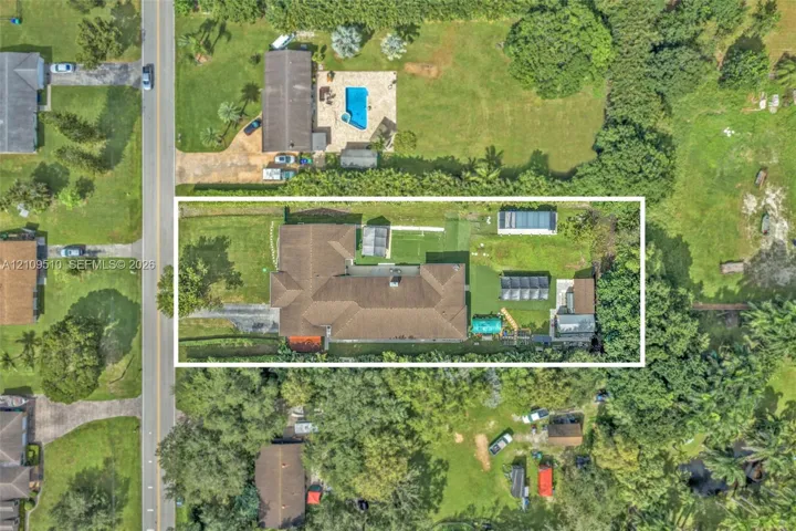 Aerial view of a residential lot featuring a multi-gabled roofline, a paved driveway, and expansive lawn space with mature trees