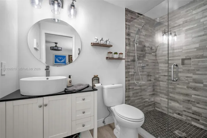 Bathroom featuring a white vanity with a dark countertop, a vessel sink, an oval wall-mounted mirror, and a walk-in shower with textured tile surround and a frameless glass door