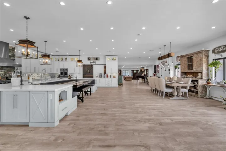 Open-concept space featuring wood-finish flooring, extensive recessed lighting, white shaker-style cabinetry, a large kitchen island with quartz countertops, and three oversized metal-frame pendant lights