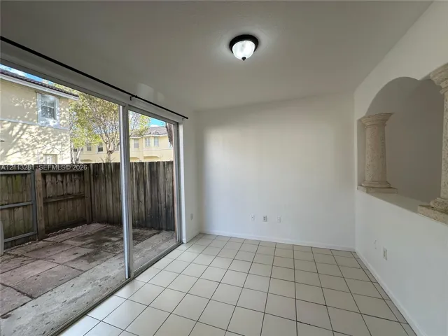 Interior space featuring light-colored tile flooring, decorative architectural columns, a ceiling-mounted light fixture, and a large sliding glass door opening to a concrete patio enclosed by wood fencing