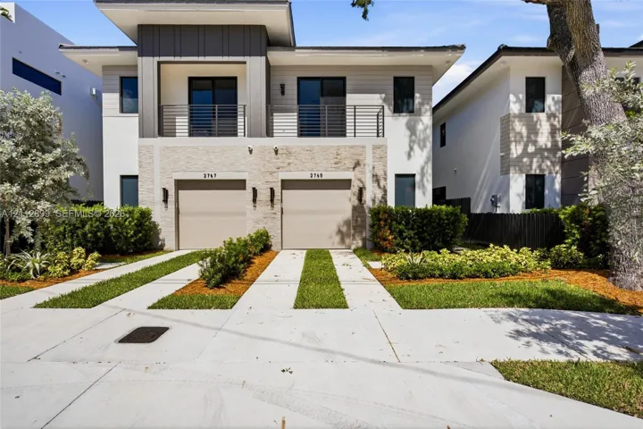 Modern exterior featuring a stacked stone facade, metal-accented second-story balcony with railings, dual two-car garage doors, and concrete paver driveways with integrated grass strips