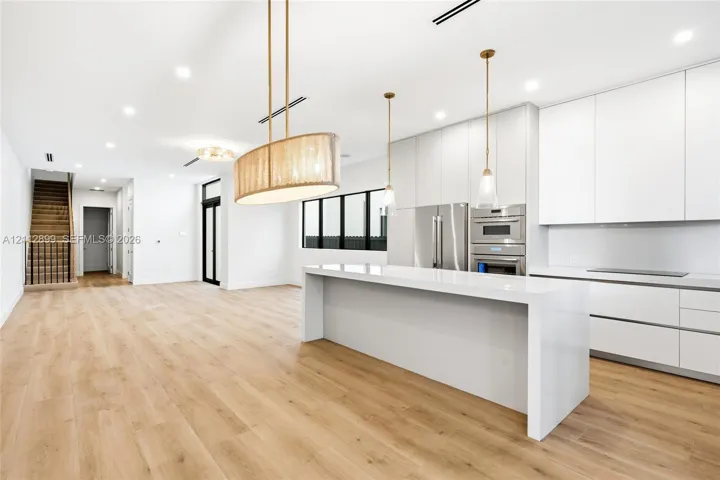 Modern kitchen featuring flat-panel cabinetry, a waterfall-edge quartz island, stainless steel built-in appliances, decorative pendant lighting, and wood-finish flooring