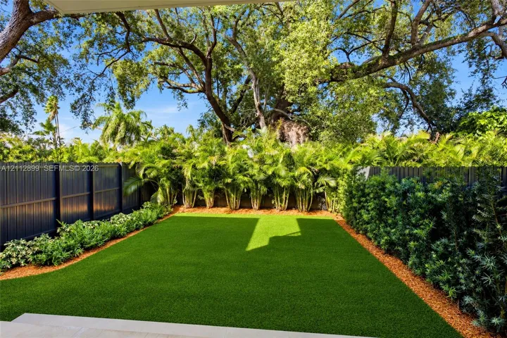 Backyard featuring artificial turf, perimeter fencing, mature canopy trees, and dense foliage border