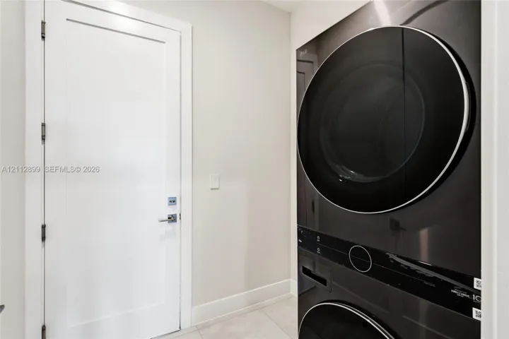 Stacked washer and dryer unit with a sleek black finish, paired with a white Shaker-style door featuring polished chrome hardware and large-format tile flooring