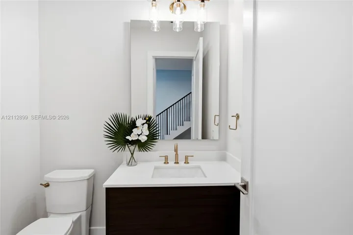 Bathroom featuring a dark wood-finish vanity, white countertop with an integrated basin, wall-mounted rectangular mirror, three-light fixture with cylindrical glass shades, and gold-tone hardware