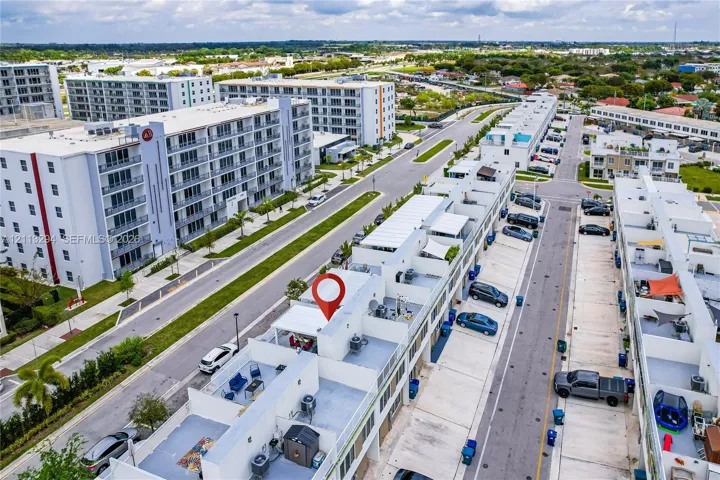 Aerial perspective of a multi-level townhome featuring a rooftop terrace, concrete construction, paved parking surface, and individual exterior utility units