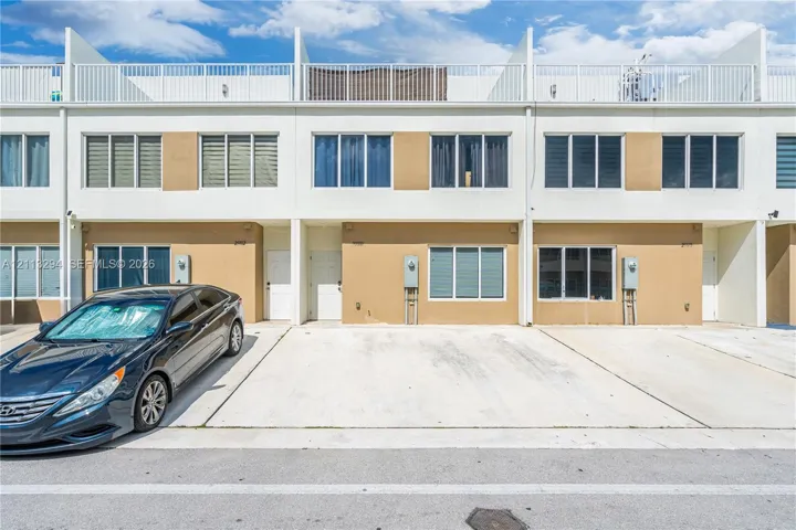 Multi-level townhome exterior featuring a smooth stucco facade, concrete driveway, and a top-level rooftop terrace with metal railings