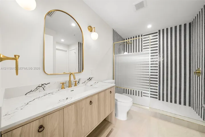 Vanity with wood-finish cabinetry, stone countertop with veining, arched wall mirror, gold-toned fixtures, and a shower enclosure featuring vertical striped mosaic tiling