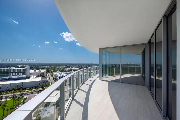 Expansive wraparound balcony featuring sleek glass railings, overhead recessed lighting, and large-format tile flooring, complemented by floor-to-ceiling glass sliding doors