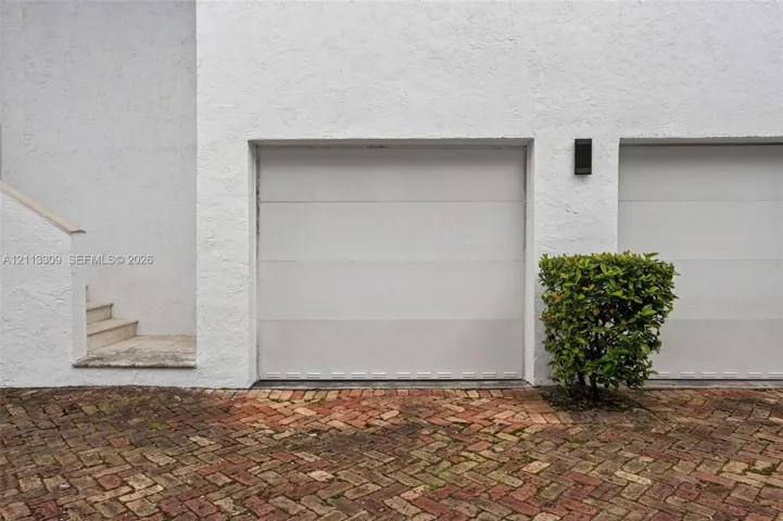 Multi-car garage featuring white paneled doors, textured stucco exterior, exterior wall-mounted lighting, and a herringbone-patterned brick paver driveway