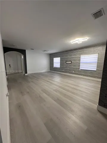 Open interior space featuring light-toned wood-finish flooring, a textured accent wall, integrated flush-mount ceiling lighting, and a prominent arched entryway